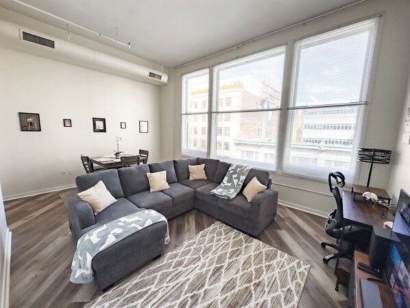 A modern living room with a sectional sofa, a dining table with chairs, and large windows with blinds. - Indy Apt, Skywalk Access, Gym & Private Parking (Indianapolis)