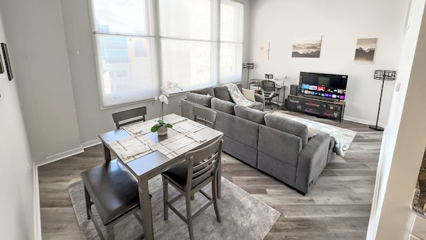 Uma sala de estar moderna com um sofá cinzento, uma mesa de jantar com cadeiras e uma televisão.