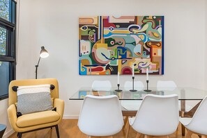 Een moderne eetruimte met een glazen tafel, witte stoelen, een gele fauteuil en een kleurrijk abstract schilderij aan de muur.