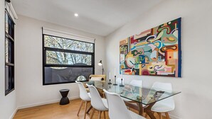 Un comedor con mesa de vidrio y sillas blancas, un cuadro abstracto grande en la pared, y una ventana con vista a árboles.