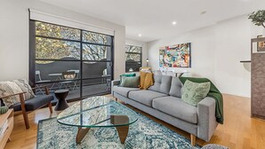 Una sala de estar moderna con una mesa de centro de vidrio, un sofá gris y ventanas grandes con vista a árboles.