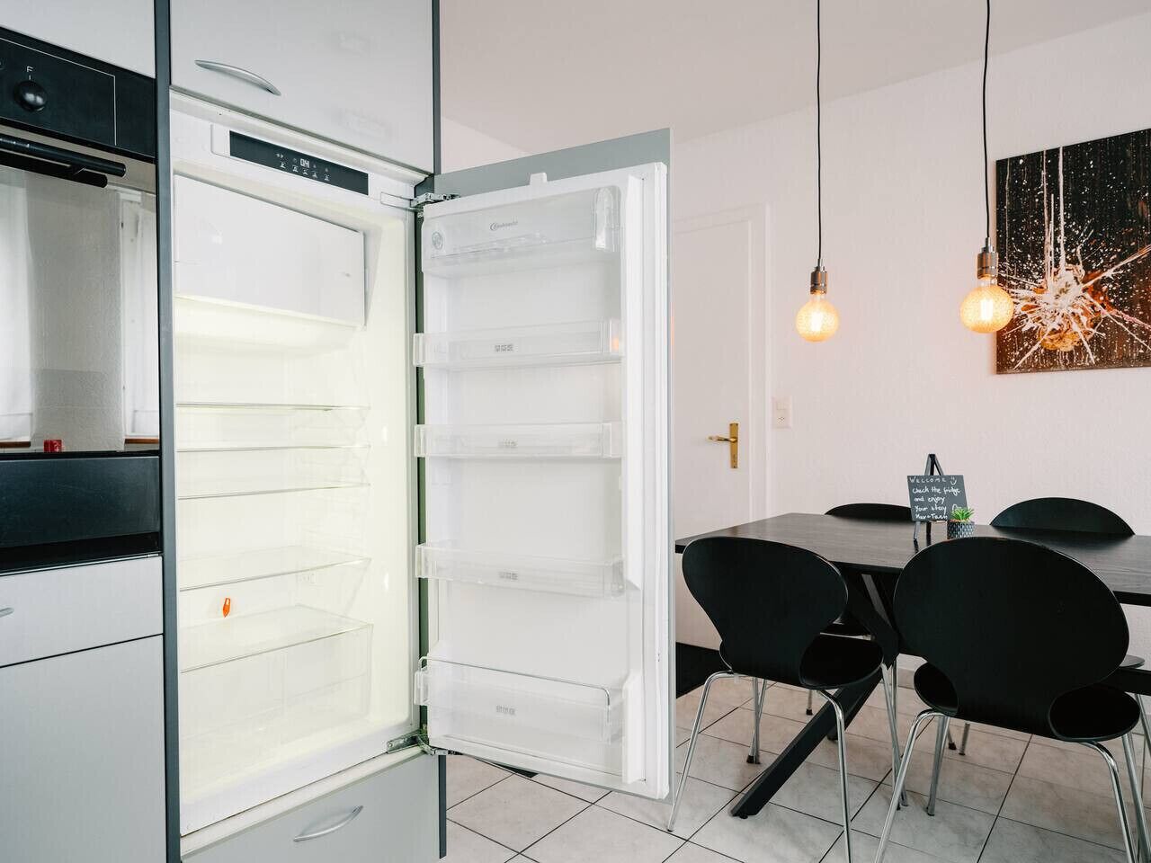 Una cocina moderna con un refrigerador blanco, contenedores de almacenamiento transparentes, una mesa de comedor con sillas negras y una lámpara de pared.