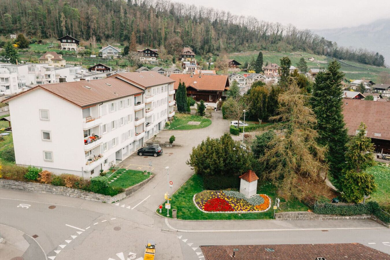 A residential area with a white multi-story building, a small garden with a flower bed, and a mountain backdrop.