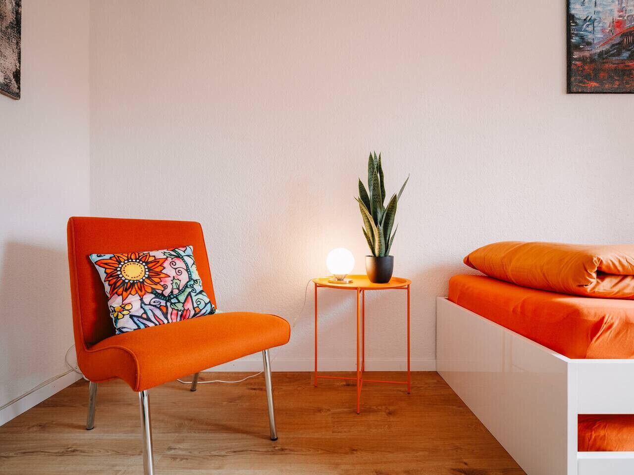 Ein Zimmer mit Holzboden, einem weißen Bett mit orangefarbener Bettwäsche, einem orangefarbenen Sessel mit farbigem Kissen und einem kleinen Tisch mit Topfpflanze und Lampe.