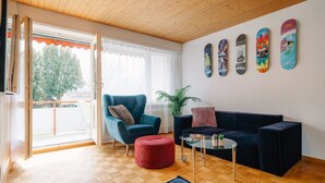 Een moderne woonkamer met een houten plafond, een blauwe fauteuil, een zwarte bank, een rode poef en een glazen salontafel.