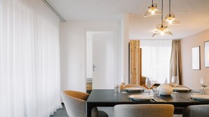 Ein Essbereich mit einem dunklen Holztisch, gedeckt für vier Personen, umgeben von gepolsterten Stühlen. Drei Pendelleuchten hängen über dem Tisch, und der Raum ist modern und minimalistisch gestaltet, mit weißen Wänden und einem großen Fenster mit Vorhängen.