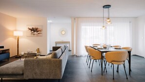 Ein modernes Wohnzimmer mit einer Couch, einem Esstisch für vier Personen und einem großen Fenster mit leichten Vorhängen.