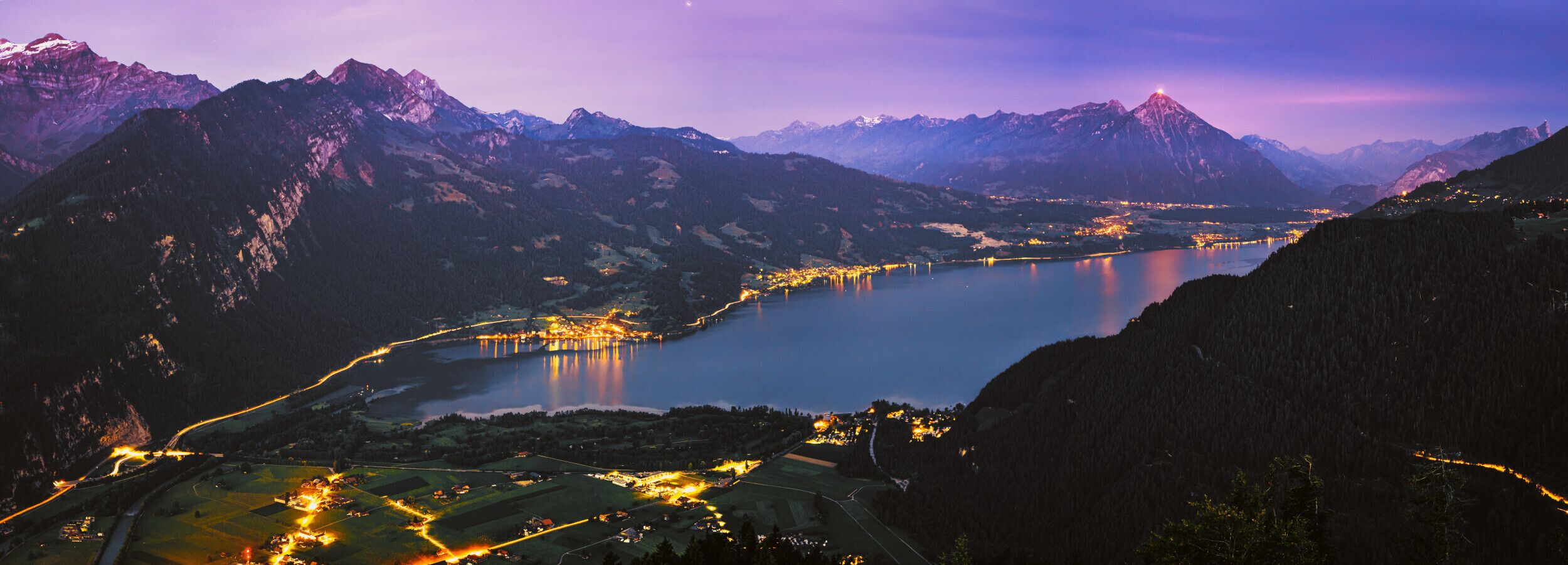 Paisagem montanhosa com um lago, estradas iluminadas e edifícios.