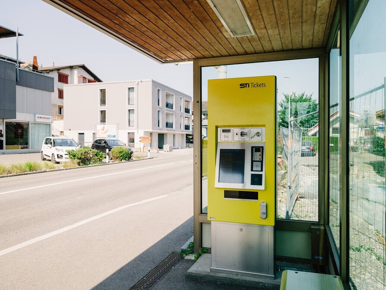 A yellow ticket vending machine at a bus stop with a wooden roof and a view of a street with cars and buildings.