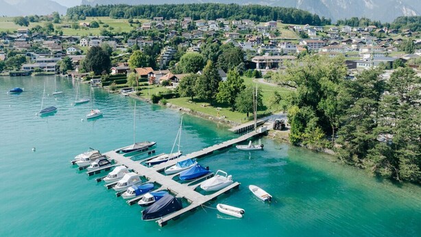 Vista aérea de un pueblo a orillas de un lago, con un puerto deportivo, casas y montañas al fondo.