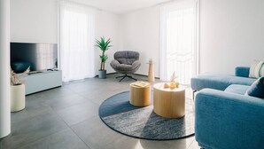 Una sala de estar moderna con un sofá gris, un sofá azul, un sillón gris, una mesa de centro redonda y un televisor sobre un mueble blanco.
