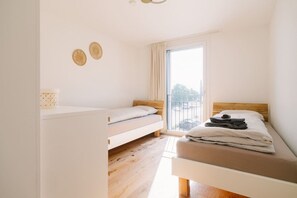 Un dormitorio con dos camas individuales, suelo de madera, un ventanal con cortinas y una mesita con una caja.
