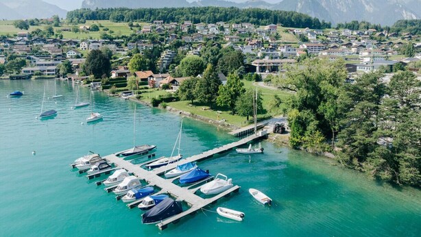 Vista aérea de un pueblo a orillas de un lago, con un puerto deportivo, casas y montañas al fondo.