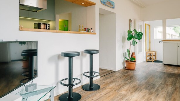 A modern kitchen with a wooden ceiling, a built-in microwave, and a TV on a glass table. There are two black bar stools and a potted plant. - Loftwohnung in Seenähe (Spiez)