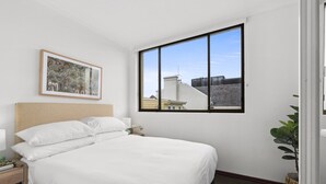 Un lit bien fait, avec du linge de lit blanc, un tableau encadré au mur et une grande fenêtre donnant sur des immeubles et un ciel dégagé.