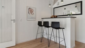 Une cuisine moderne avec un mur recouvert de panneaux en bois blancs, un plan de travail noir, trois tabourets de bar noirs, un luminaire suspendu et une œuvre d’art encadrée au-dessus du plan de travail.