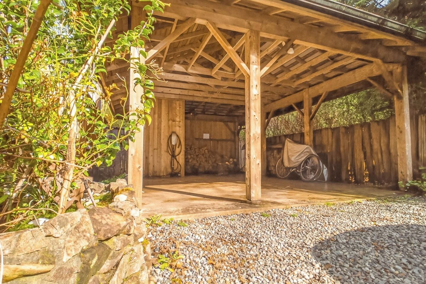 Un espacio exterior techado con estructura de madera, suelo de grava y una silla de ruedas.
