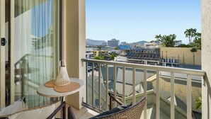 A balcony with a glass door, a wicker chair, a small table with a woven placemat, and a view of a cityscape with buildings and palm trees. - Luxurious City Centre Studio - Space & Style (Cairns City)