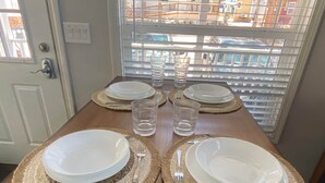 Una mesa de comedor dispuesta con platos, cubiertos y vasos, ubicada frente a una ventana con persianas.