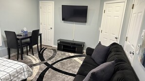 A compact living room with a black sofa, a small TV, a dining table with chairs, and a patterned rug.