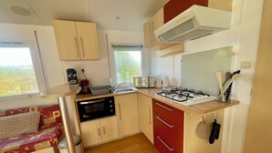 Une cuisine compacte équipée d’un four à micro-ondes, d’un grille-pain et d’une cuisinière à gaz. Une fenêtre laisse entrer la lumière naturelle, et un petit coin salon avec un coussin est à disposition.