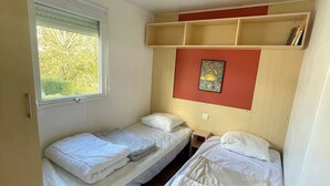 Une petite pièce, de taille modeste, comprenant deux lits simples, une fenêtre avec des stores, un luminaire au plafond et une étagère remplie de livres.