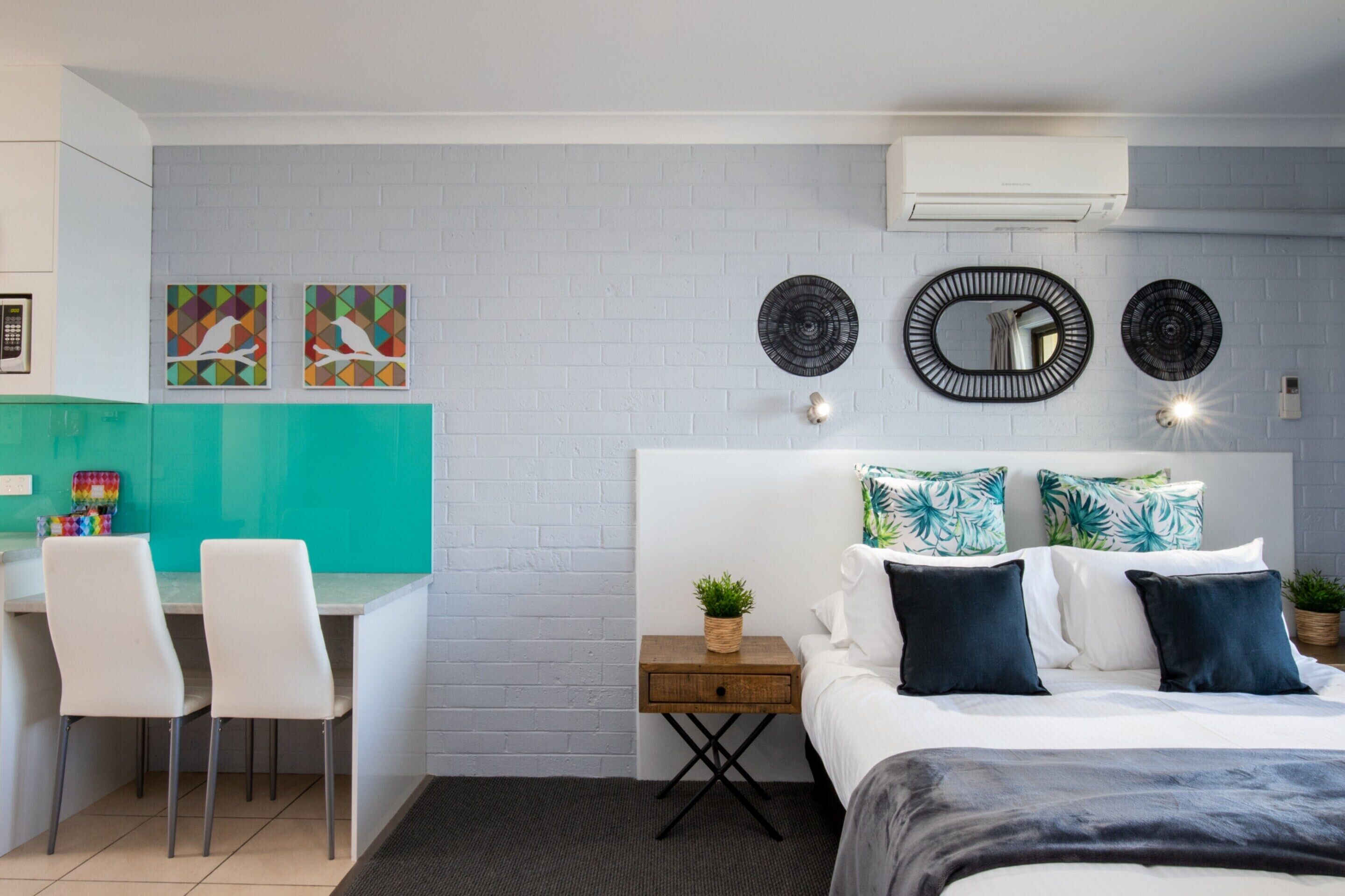 A modern hotel room with a white brick wall, a bed with dark bedding, a small bedside table with a plant, and a desk with chairs.