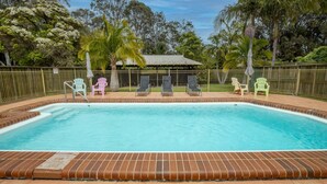 A swimming pool with lounge chairs and a covered seating area, surrounded by a fence and trees. - Batemans Bay Studio Escape With Lush Vista (Batehaven)