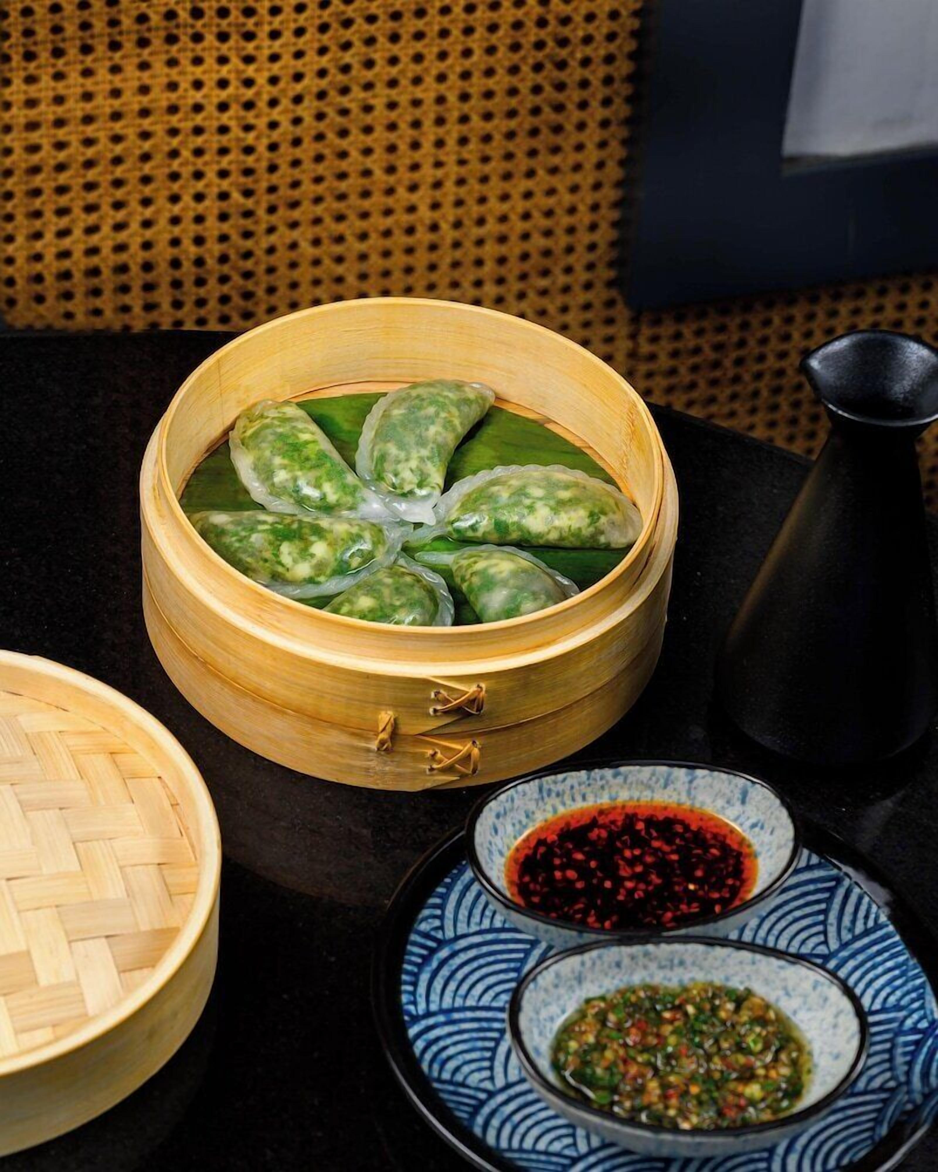 A bamboo steamer with dumplings, a woven basket, and two bowls of dipping sauces.