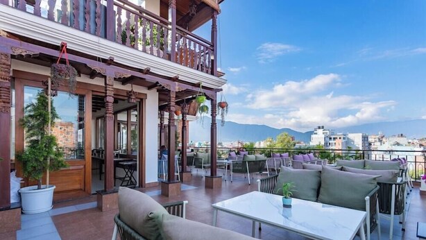 An outdoor seating area with tables and chairs, a balcony with hanging plants, and a view of the cityscape. - Timber House (Kathmandu)