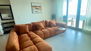 Uma sala de estar ampla, com um sofá seccional grande, uma mesa redonda pequena e uma janela grande que oferece uma vista.