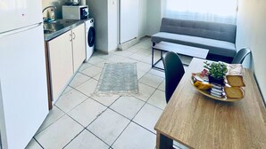 Eine kleine Küche mit Waschmaschine, einem Esstisch mit Pflanze und Brot, sowie ein graues Sofa im Hintergrund.