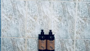 Zwei Flaschen Olivenöl-Shampoo und Flüssigseife auf einer gefliesten Wand.