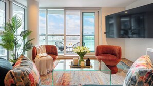 Ein modernes Wohnzimmer mit einem Glastisch, zwei roten Sesseln, einem schwarzen Sofa, einem großen Fernseher und Blick auf die Stadt durch große Fenster.