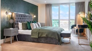 Ein modernes Schlafzimmer mit einem gesteppten Kopfteil, einem großen Bett mit einer grünen Steudecke, einem Nachttisch mit einer Dekoration, einem Schreibtisch mit einer Lampe und Blick ins Freie durch ein großes Fenster.