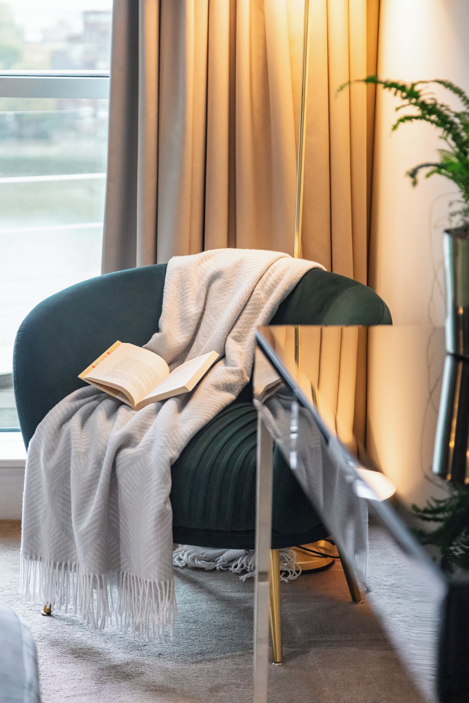 Un salón moderno con un sillón verde oscuro, una manta blanca y un libro abierto. Se ve una lámpara de pie y una planta en maceta al fondo.