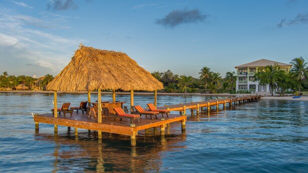 A wooden pier with lounge chairs extending over calm water, a thatched-roof structure, and a multi-story building with balconies in the background.