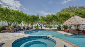 A swimming pool with lounge chairs and a thatched umbrella, surrounded by trees and a beach view.