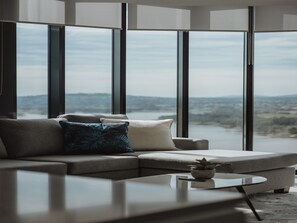 A modern living room with a sofa, a coffee table, and a view of a body of water through large windows. - Panoramic Lake View 2-bed With Pool, (Canberra)