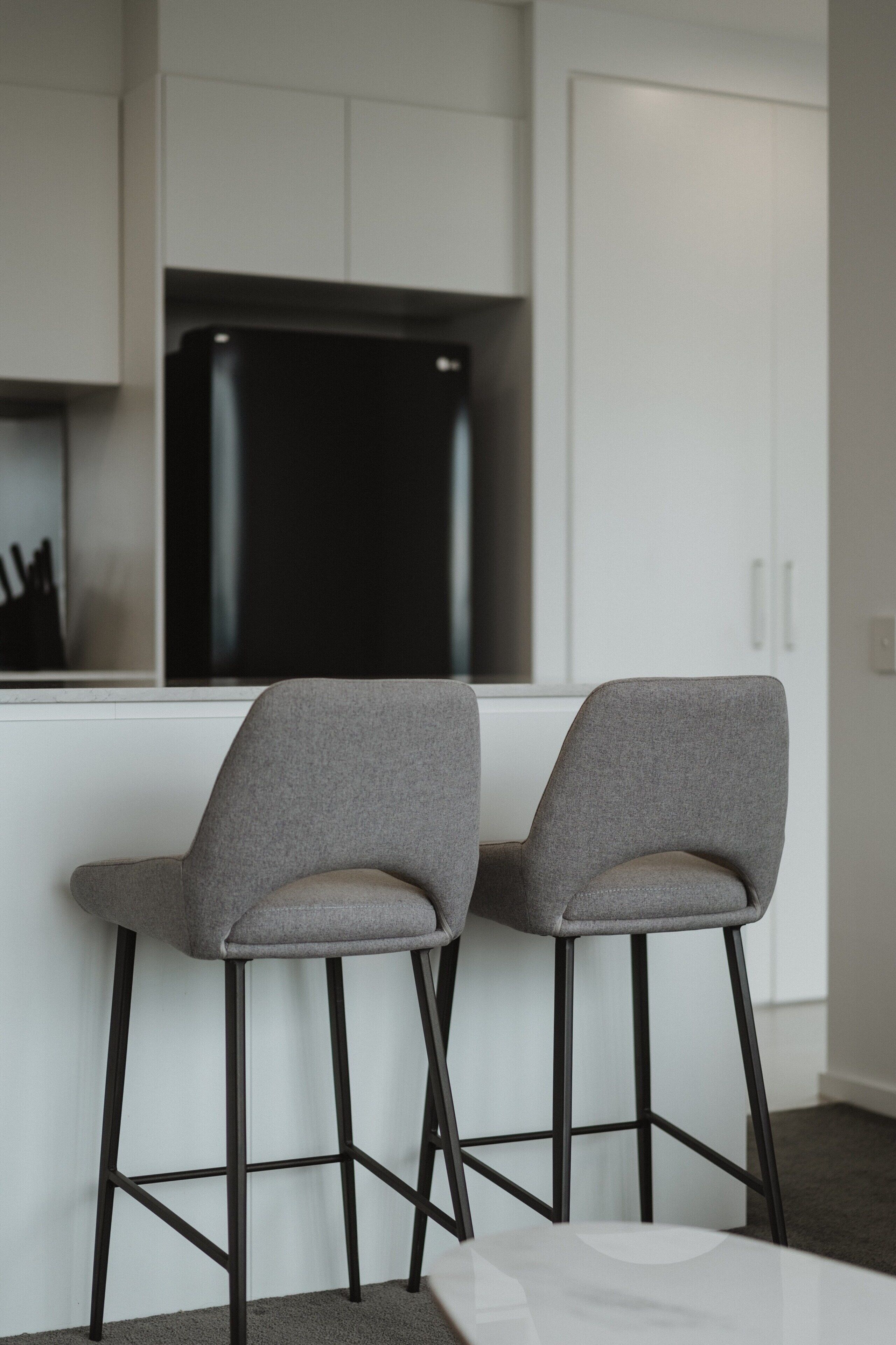 A modern kitchen with two grey bar stools, a black refrigerator, and white cabinetry.