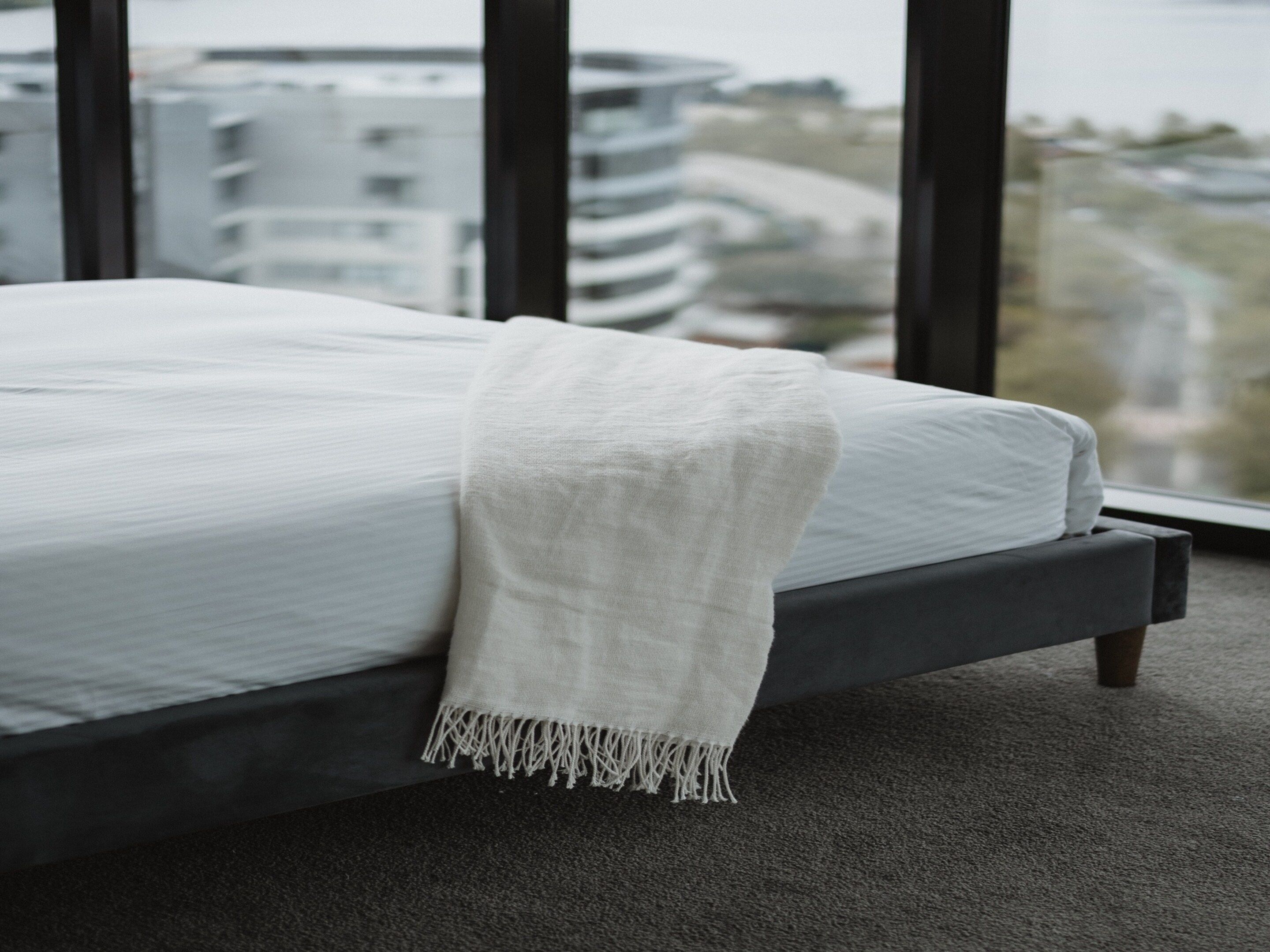 A bed with a white blanket and a view of buildings through large windows.