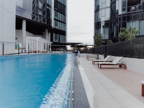 A swimming pool with lounge chairs and a covered seating area, surrounded by modern buildings. - Panoramic Lake View 2-bed With Pool, (Canberra)