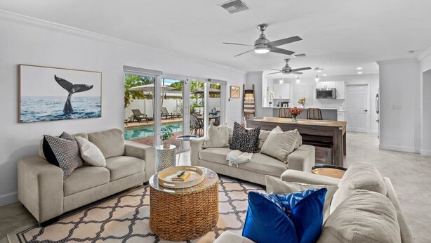 A spacious living room with a large window overlooking a pool, a kitchen area, and a ceiling fan. - Stylish 4BR Oasis | Heated Pool, Spa & Fire Pit (Pompano Beach)