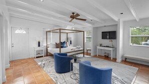 A spacious bedroom with a four-poster bed, a ceiling fan, a TV, and a large window with blinds. - Stylish 4BR Oasis | Heated Pool, Spa & Fire Pit (Pompano Beach)