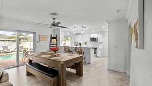 A modern kitchen with white cabinetry, a dining area with a wooden table, and a pool visible through the glass doors. - Stylish 4BR Oasis | Heated Pool, Spa & Fire Pit (Pompano Beach)