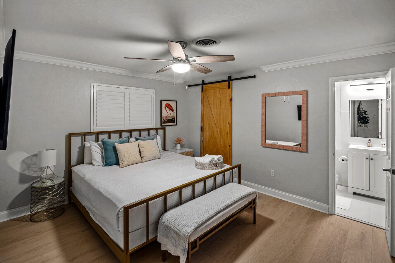A bedroom with a bed, bedside lamp, wooden floor, ceiling fan, and a door leading to a bathroom.