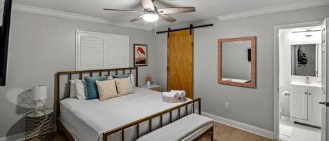 A bedroom with a bed, bedside lamp, wooden floor, ceiling fan, and a door leading to a bathroom.
