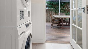 Un espace buanderie équipé d’un lave-linge et d’un sèche-linge à chargement frontal, d’une table en bois avec des chaises, et d’une porte vitrée donnant sur un patio extérieur avec vue sur la verdure.
