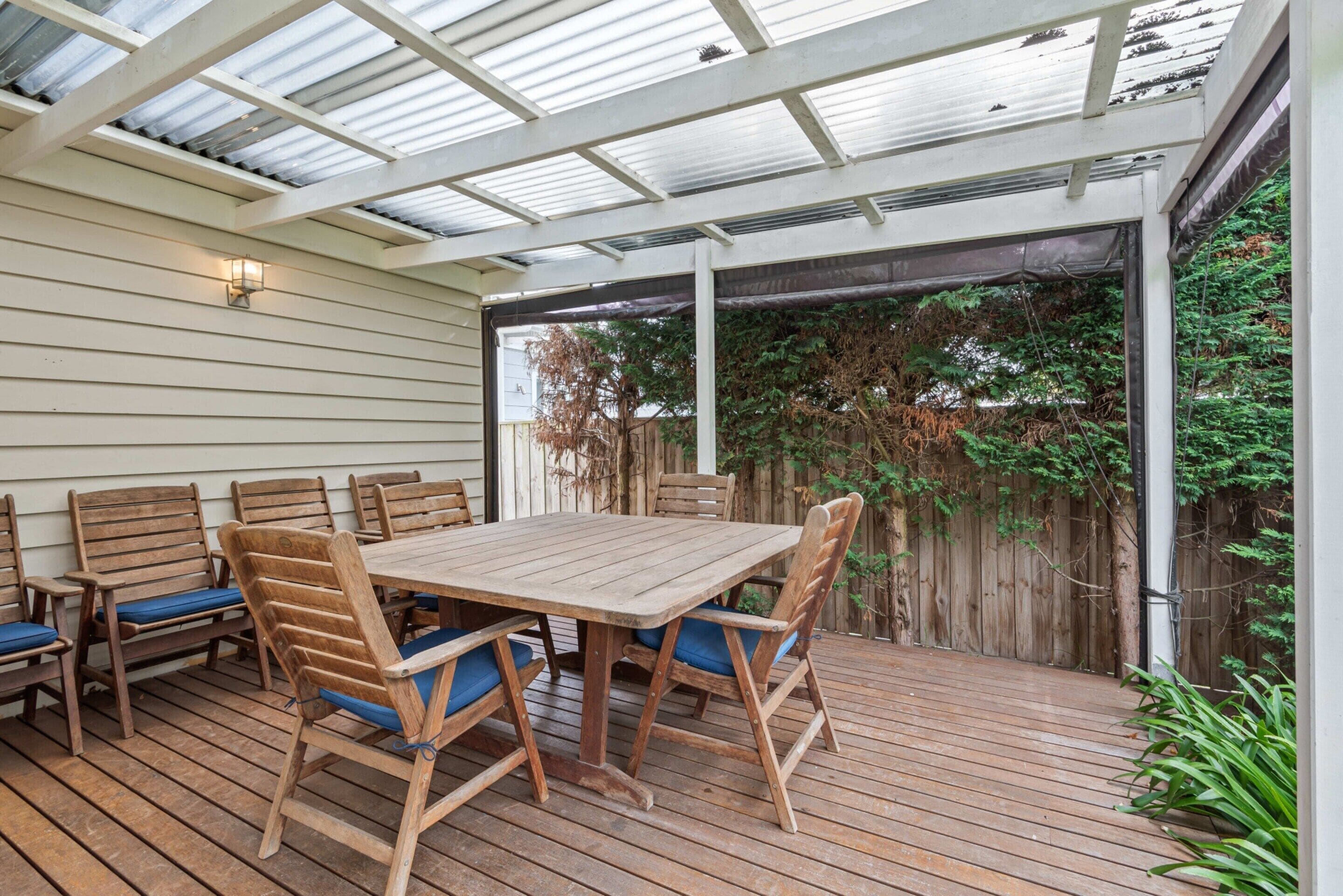 An outdoor patio with a wooden table and chairs, a pergola with a glass roof, and a wooden fence in the background.