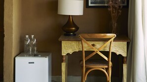 Una habitación con un escritorio de madera, una mesita de noche blanca, una lámpara, una silla, un cuadro enmarco y un florero con flores secas.